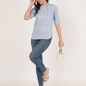 May include: A light blue linen top with a button-up closure on the side. The top has a mandarin collar and three-quarter sleeves. The model is wearing blue jeans and white heeled sandals.