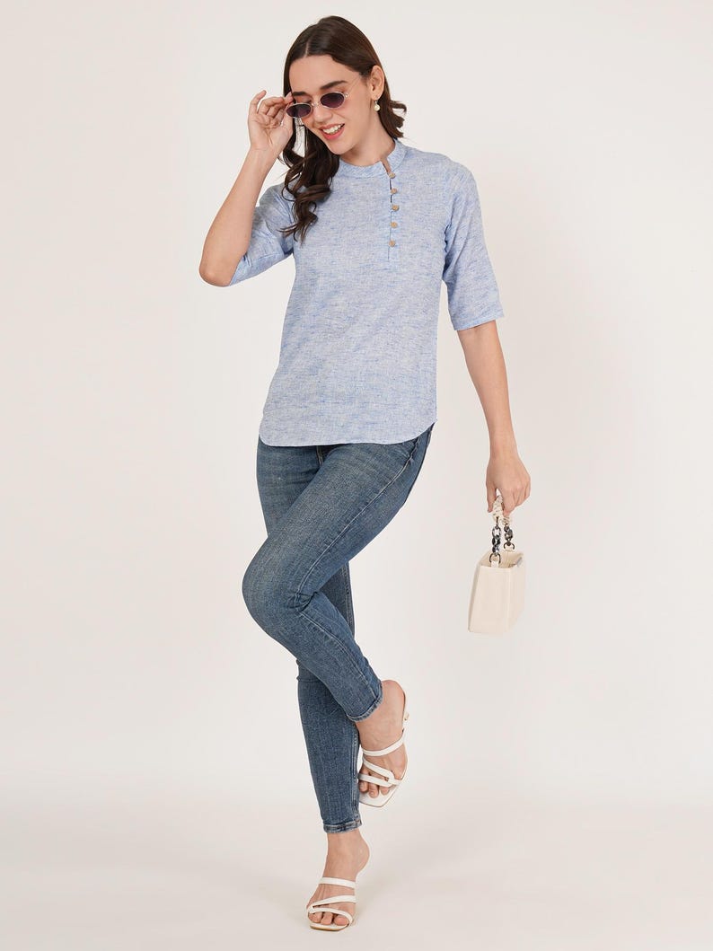 May include: A light blue linen top with a button-up closure on the side. The top has a mandarin collar and three-quarter sleeves. The model is wearing blue jeans and white heeled sandals.
