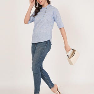 May include: A woman wearing a light blue, textured, button-down top with three-quarter sleeves and blue jeans. She is holding a white handbag with a chain strap and wearing white heeled sandals.