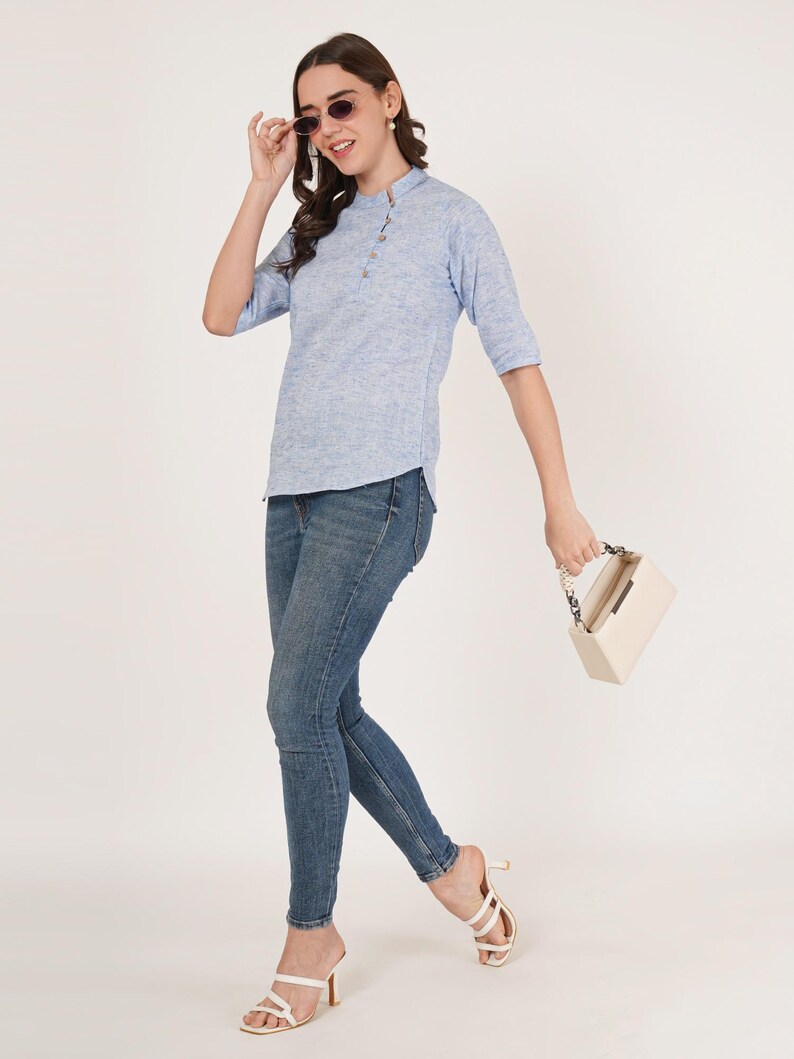 May include: A woman wearing a light blue, textured, button-down top with three-quarter sleeves and blue jeans. She is holding a white handbag with a chain strap and wearing white heeled sandals.