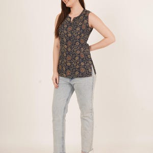 May include: Sleeveless navy top with a floral pattern in gold and orange. The top features a notched neckline and a side slit. The model is wearing light wash denim jeans and clear strap heels.