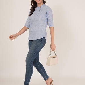 May include: A woman wearing a light blue, textured, collared shirt with a button placket and three-quarter sleeves. She is wearing blue jeans and white heeled sandals. She is carrying a white handbag with a chain strap.