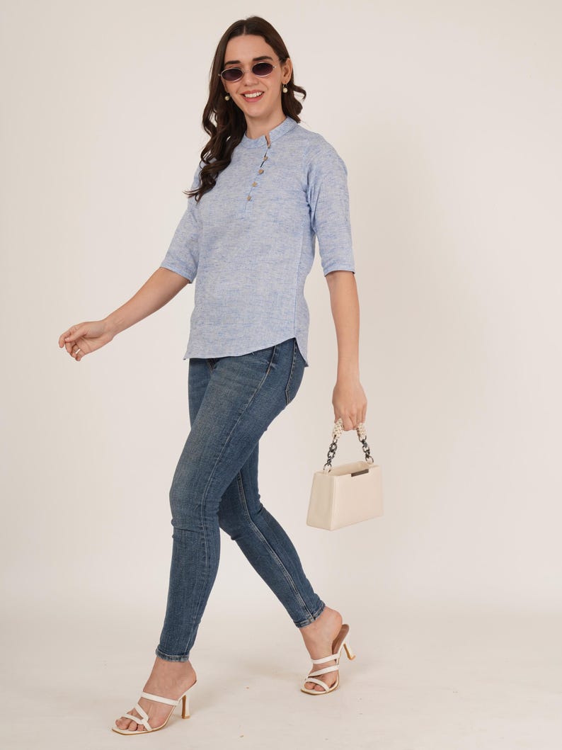May include: A woman wearing a light blue, textured, collared shirt with a button placket and three-quarter sleeves. She is wearing blue jeans and white heeled sandals. She is carrying a white handbag with a chain strap.