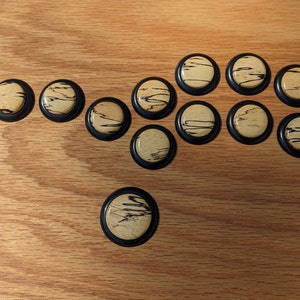 May include: A collection of twelve round wooden cabinet knobs with a black border. The knobs have a light tan wood grain pattern with dark brown streaks. They are arranged on a light brown wooden surface.