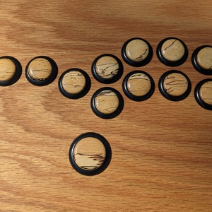 May include: A collection of twelve round wooden game pieces with a black border. The wooden centers have a light tan color with dark brown grain patterns. The pieces are arranged on a light brown wooden surface.