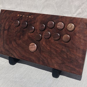 Może przedstawiać: Ręcznie wykonany drewniany kontroler do gier arkadowych o bogatym, ciemnobrązowym wykończeniu. Posiada szereg okrągłych przycisków w różnych odcieniach brązu oraz zestaw mniejszych przycisków wzdłuż górnej części, osadzonych w drewnianej powierzchni.