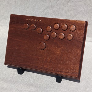 Może przedstawiać: Drewniany kontroler do gier arkadowych o bogatym, ciemnobrązowym wykończeniu. Posiada szereg okrągłych przycisków i układ joysticka, przeznaczony do gier. Kontroler jest podparty dwoma czarnymi nóżkami.