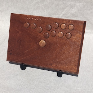 Może przedstawiać: Drewniany kontroler do gier arcade w kolorze ciemnobrązowym. Posiada szereg okrągłych przycisków w klasycznym układzie, idealnym do gier. Kontroler jest podparty dwoma czarnymi nóżkami.