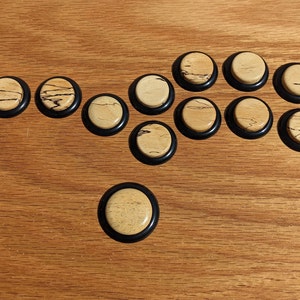 May include: A collection of wooden game pieces with a light wood grain center and a black rubber ring around the edge. The pieces are arranged on a wooden surface, showcasing their contrasting colors and textures. The game pieces are round.
