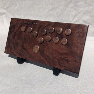 Może przedstawiać: Drewniany kontroler do gier arkadowych w głębokim brązowym odcieniu. Posiada konfigurację przycisków i joysticka, idealną do gier. Kontroler jest podparty dwoma czarnymi nóżkami, lekko go podnosząc. Widoczne usłojenie drewna.