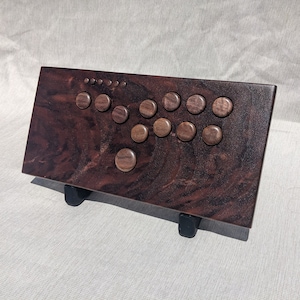 Może przedstawiać: Niestandardowy kontroler do gier arkadowych z ciemnobrązowym wykończeniem z usłojeniem drewna. Posiada drewniane przyciski i układ joysticka. Kontroler jest zamontowany na czarnym stojaku. Idealny do gier retro.