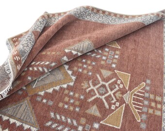 Op de afbeelding: Een opgevouwen, roodbruin tapijt met geometrische patronen in tinten grijs, beige en wit. Het tapijt heeft een gedetailleerde rand en een franje langs één rand. Het ontwerp omvat driehoeken, vierkanten en gestileerde vormen.