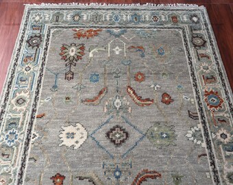 May include: A rectangular, gray area rug with an intricate, floral pattern. The rug features a central field with repeating motifs in shades of blue, orange, and cream. The border is a darker gray with a detailed design. The rug is made of a soft material.