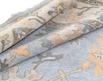 May include: A rolled-up, light blue rug with a detailed floral pattern in shades of peach, beige, and green. The rug has a textured appearance and a fringed edge, showcasing a traditional design.