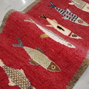 Puede incluir: Una alfombra rectangular roja con un diseño de peces. La alfombra presenta múltiples peces en varios colores, incluyendo beige, blanco y negro, sobre un fondo rojo. La alfombra tiene un borde beige.