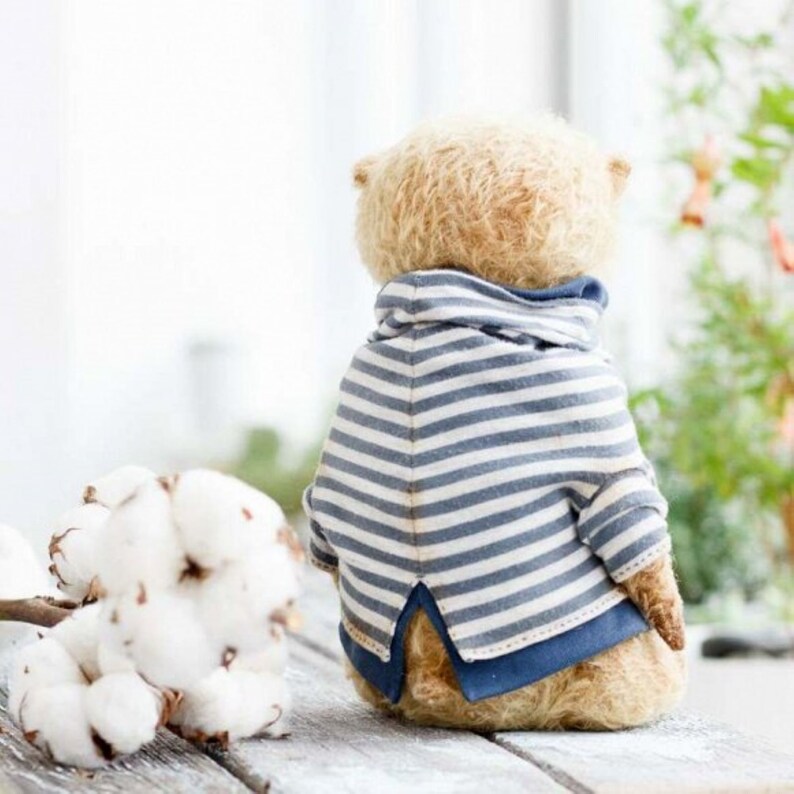 Op de afbeelding: Een beige teddybeer die een blauw-wit gestreept shirt draagt met een blauwe kraag. De beer zit op een houten oppervlak met een bos witte wattenbolletjes op de voorgrond.