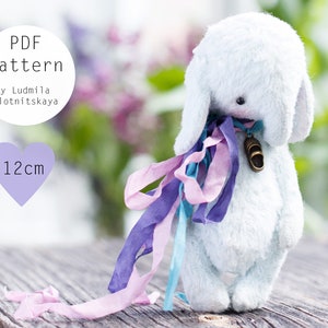 Puede incluir: Un juguete de peluche de conejo azul claro con orejas largas y una cinta alrededor del cuello. El conejo tiene un pequeño dije de bronce y cintas de colores. La imagen incluye el texto "PDF pattern" y "12cm".