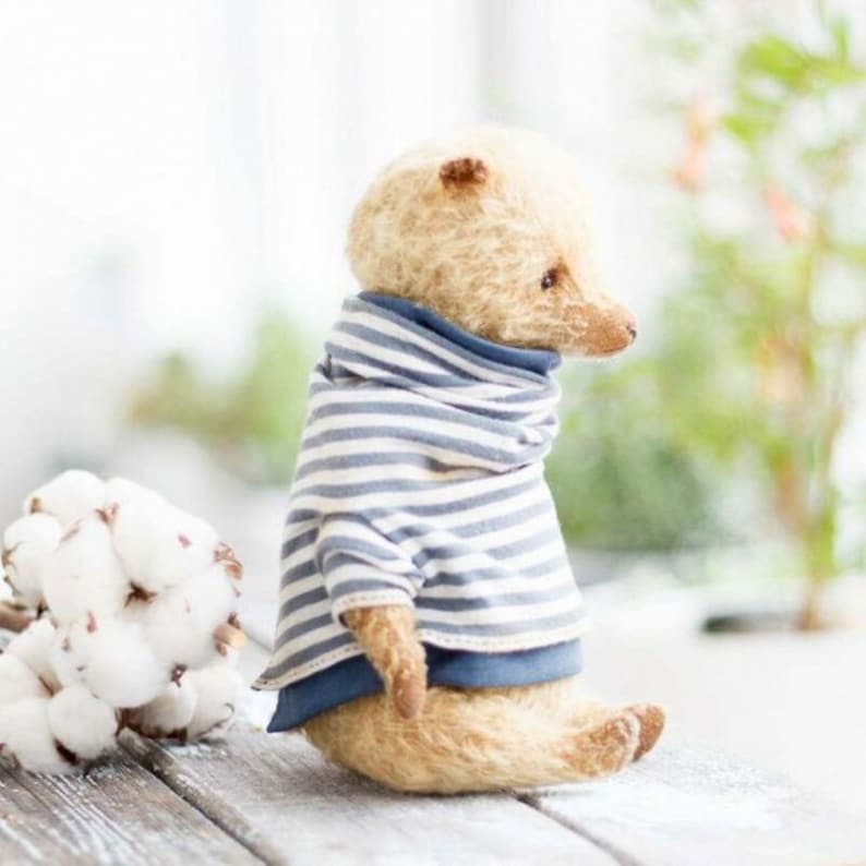 Op de afbeelding: Een kleine, cr&egrave;mekleurige teddybeer die een blauw-wit gestreept shirt draagt met een blauwe turtleneck. De beer zit op een licht houten oppervlak met een witte katoenen bol in de achtergrond.