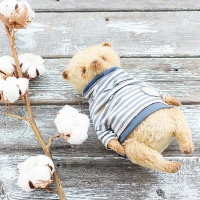 Op de afbeelding: Een lichtbruine teddybeer die een blauw-wit gestreept shirt draagt met een blauwe kraag. De beer ligt op een houten oppervlak met een katoenen tak op de achtergrond.