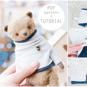 May include: A brown teddy bear wearing a white sweater with blue trim and a pair of brown buttons. The teddy bear is being held by a hand. The image includes the text "PDF pattern & TUTORIAL" and "50 photos".
