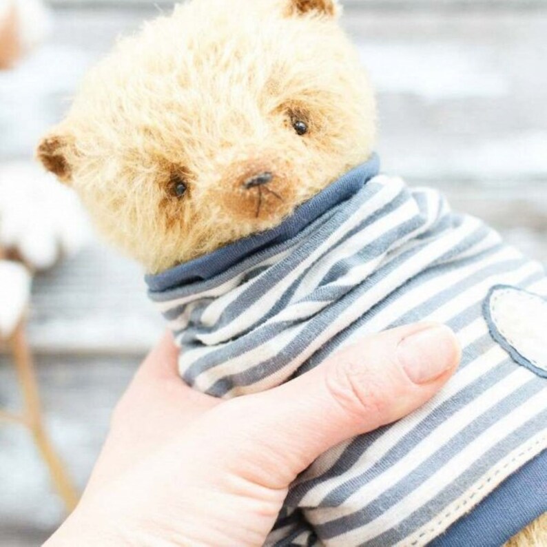 Op de afbeelding: Een lichtbruine teddybeer die een blauw-wit gestreept shirt draagt met een wit hartje op de voorkant.