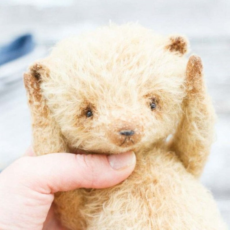 Op de afbeelding: Een kleine, cr&egrave;mekleurige teddybeer met een pluche, ruwe vacht. De beer heeft bruine ogen en een kleine, zwarte neus. De beer wordt vastgehouden door een hand.