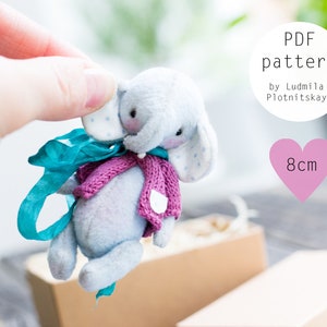 May include: A PDF pattern for a small, gray elephant plush toy with a pink knitted scarf and a blue ribbon. The elephant is 8 cm tall. The pattern is by Ludmila Plotnitskaya.