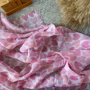 May include: A pink and white scarf with a heart pattern. The scarf features a repeating design of pink hearts in varying shades, creating a romantic and feminine aesthetic. The fabric appears lightweight and flowing, with a fringed edge.
