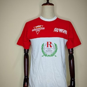 May include: Red and white short-sleeve t-shirt with racing team logos. The shirt features the text "Group C Nissan Racing Team" and "NISMO" on the front. A large emblem with the text "Racing Motorsports International" is also visible.