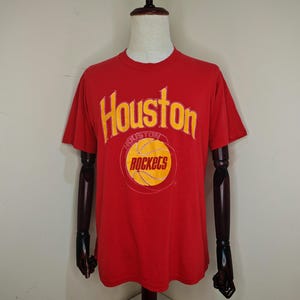 May include: Red t-shirt with the words "Houston" and "Rockets" in yellow lettering. The word "Houston" is arched above a yellow basketball graphic with the word "Rockets" inside. The shirt is displayed on a mannequin.