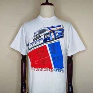 May include: White t-shirt featuring a graphic of a Porsche race car with the text "Rothmans Porsche 962C". The shirt also includes the text "Porsche-Sieg" in red and blue blocks. The shirt is displayed on a mannequin.