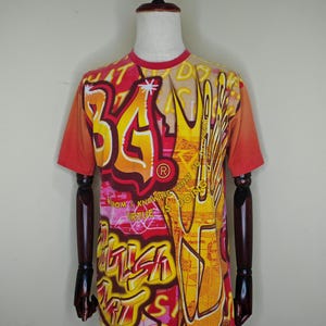 May include: A short-sleeved t-shirt with a vibrant, graffiti-style design. The shirt features a red collar and sleeves, with a colorful graphic print in shades of orange, yellow, and pink. The design includes text and a hand illustration.
