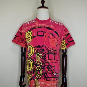 May include: Red t-shirt with a graphic design featuring the words "BODY" and "Glove" in yellow and black. The shirt has a crew neck and short sleeves. The text "EVERYBODY GLOVE BUILT FOR LIFE" is also visible.