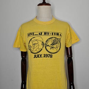 May include: A bright yellow short-sleeve t-shirt featuring a black graphic print. The text reads LIVE...AT JFK-PHILA and JULY, 1978. The graphic includes a circular illustration of a person with glasses and a winged horse, known as Pegasus.