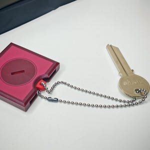 May include: A key and a key holder connected by a silver ball chain. The key holder is a square shape with a circular indentation and a keyhole. The key is a standard design. The key holder is a deep red color.