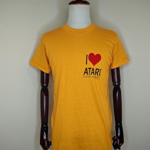 May include: Mustard yellow t-shirt with the text "I ❤️ ATARI VIDEO GAMES" in black and red. The shirt is short-sleeved and has a crew neck. The Atari logo is printed on the front.