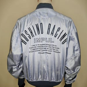 May include: A silver bomber jacket with the text "HOSHINO RACING IMPUL" printed on the back. The jacket has a gray collar and cuffs, and a quote by Kazuyoshi Hoshino is printed below the logo.