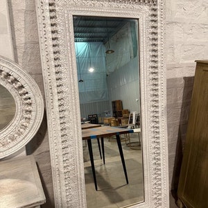 Puede incluir: Un espejo de madera blanco, tallado de forma ornamental con un marco rectangular. El espejo refleja una habitación con suelo de madera y una mesa con patas negras.