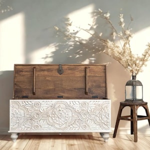 May include: A wooden chest with a white, carved floral design on the front and a dark brown top. The chest is open, revealing the interior. A small, dark brown wooden stool holds a glass lantern with a plant inside.