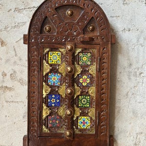 以下が含まれることがあります： アーチ型のトップと装飾的な彫刻が施された木製キャビネット。 キャビネットのドアには、真鍮のアクセントで縁取られた、花柄のカラフルな正方形のタイルが特徴です。 キャビネットは濃い茶色です。