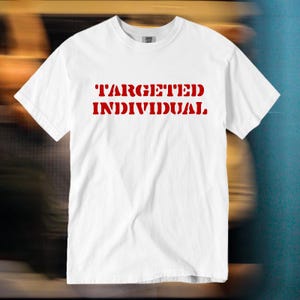 Puede incluir: Una camiseta blanca de manga corta con el texto "TARGETED INDIVIDUAL" impreso en letras mayúsculas rojas y negritas en el pecho. La prenda tiene un cuello redondo estándar y un dobladillo recto, confeccionada en tela blanca lisa.