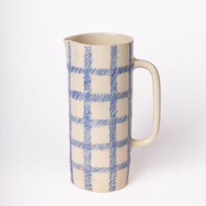 Peut inclure: Un pichet en céramique blanc cassé avec un motif à carreaux bleu. Le pichet a une grande anse et un bec verseur. Le motif à carreaux est composé de lignes bleues entrecroisées, créant un motif en forme de grille sur la surface.