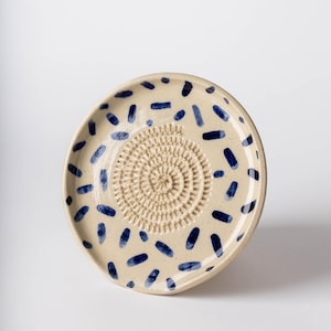 Peut inclure: Une râpe à fromage en céramique avec une base beige et une surface de râpage texturée centrale. La plaque est décorée de tirets bleu marine. Cet ustensile de cuisine est idéal pour râper les ingrédients.