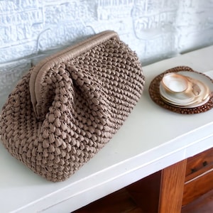 May include: A brown woven clutch bag with a textured surface. The bag is closed and has a rounded shape.