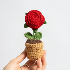 Könnte beinhalten: Eine gehäkelte rote Rose mit grünen Blättern in einem gehäkelten Topf aus Braun und Beige.