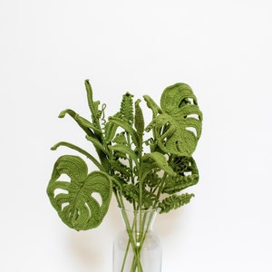 Pode incluir: Um buquê de folhas verdes de crochê em um vaso de vidro transparente. As folhas são dispostas em forma de leque e são feitas de um fio macio e texturizado.