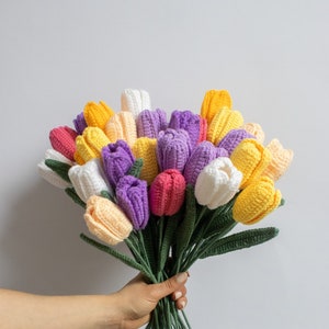 May include: A bouquet of crocheted tulips in shades of yellow, white, purple, and pink. The tulips are arranged in a hand-held bouquet with green stems.