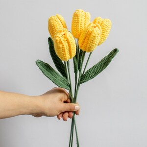 May include: A bouquet of crocheted yellow tulips with green leaves. The tulips are held in a hand.