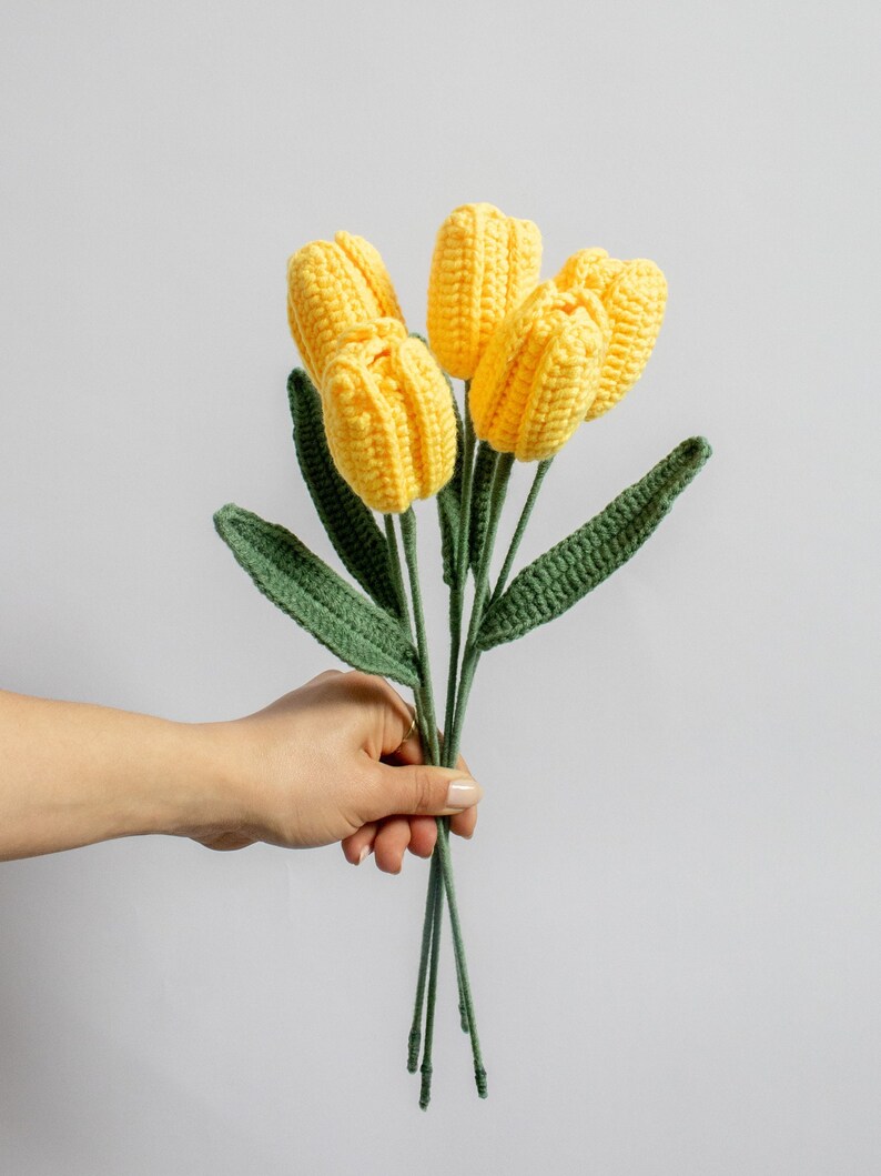 May include: A bouquet of crocheted yellow tulips with green leaves. The tulips are held in a hand.