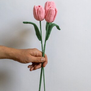 May include: Two pink crocheted tulips with green stems and leaves. The tulips are held in a hand.
